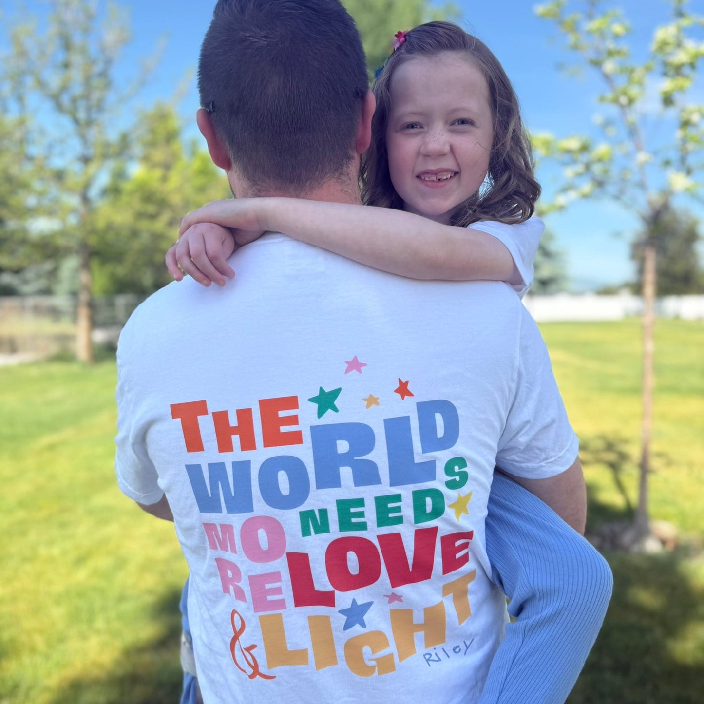 Man carrying a young girl outdoors with a colorful t-shirt message.