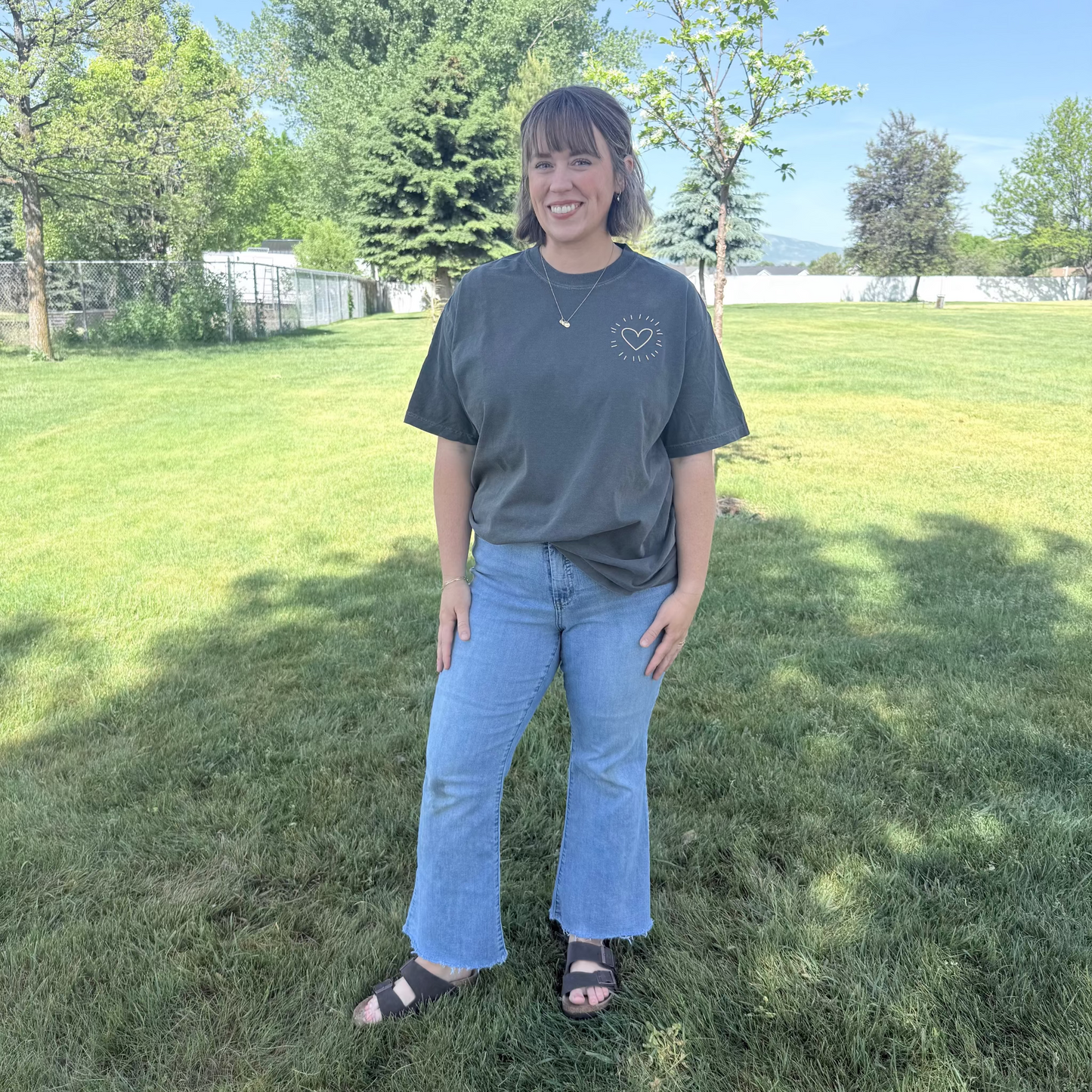 Person wearing a dark gray t-shirt and blue jeans standing on grass with trees in the background