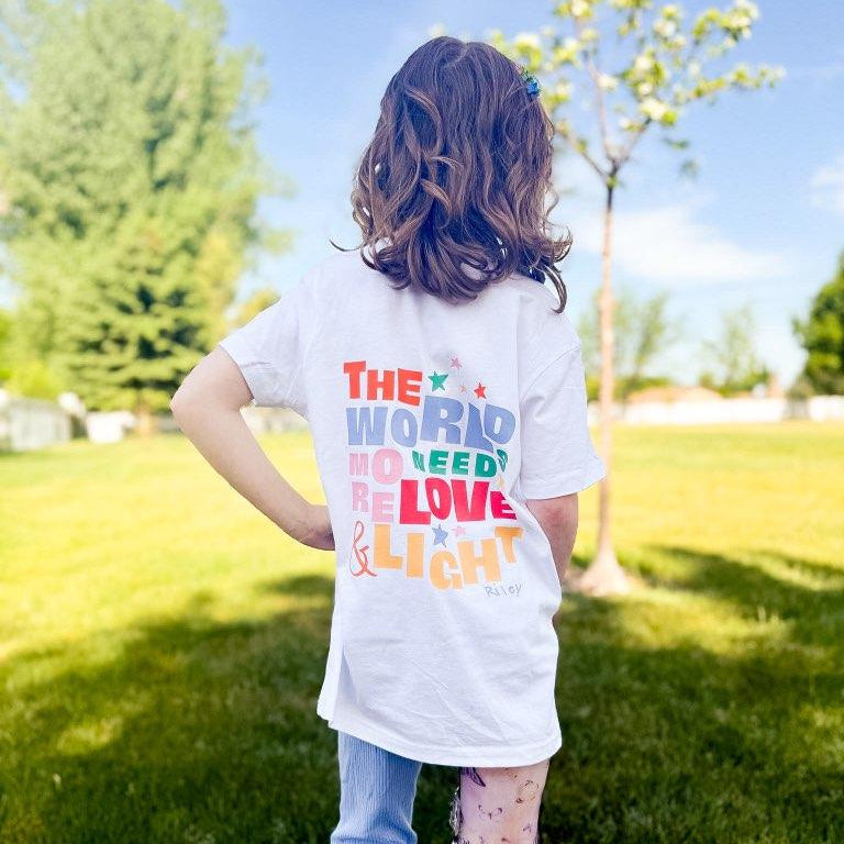 Child wearing a white t-shirt with colorful text in an outdoor setting